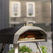 Ooni Koda 12 Outdoor Pizza Oven on a stand with a pizza baking on the stone inside and a blurred building behind.