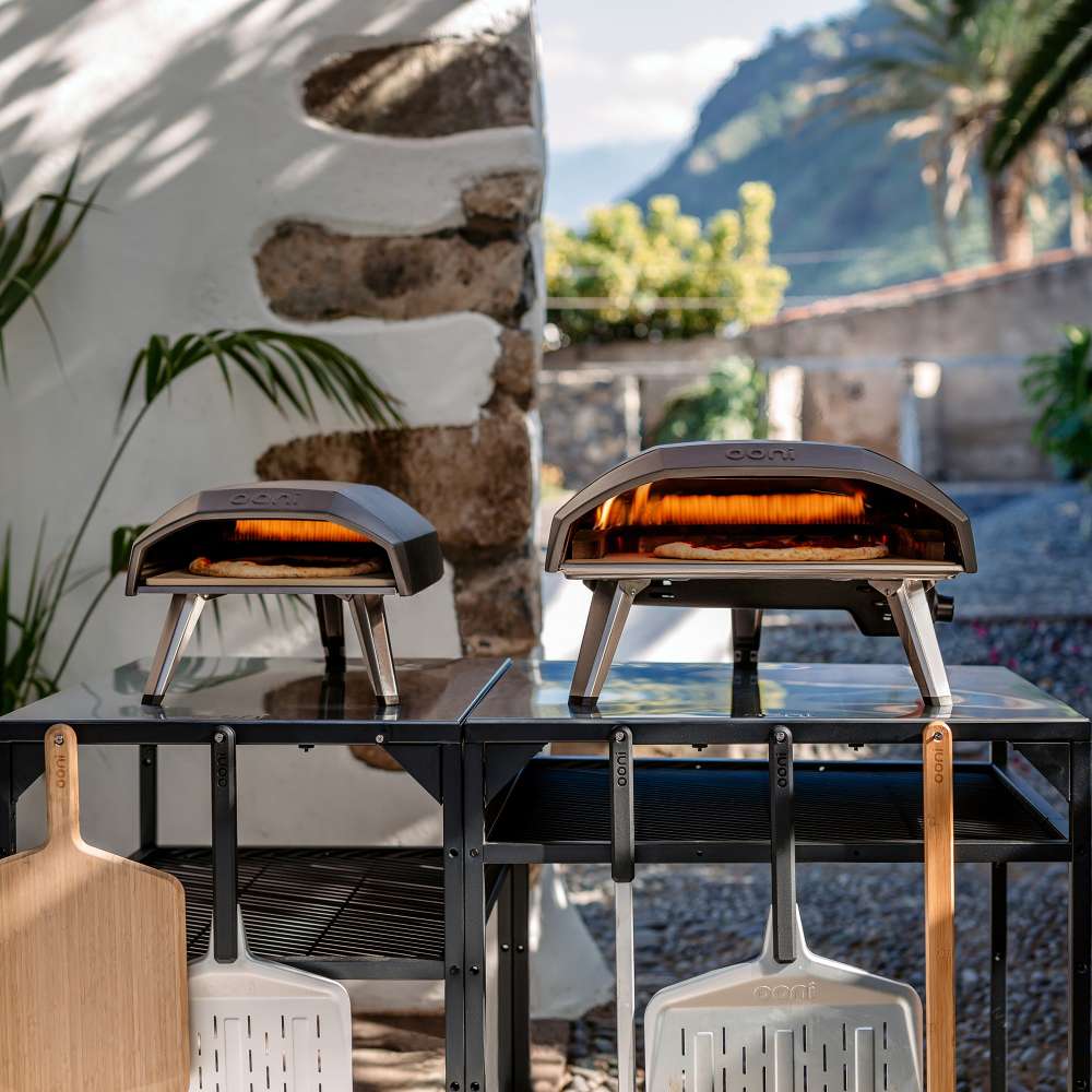 Ooni Koda 12 Outdoor Pizza Oven shown as two lit ovens side by side on a black outdoor table against a white wall with peels leaning below.