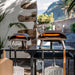 Ooni Koda 12 Outdoor Pizza Oven shown as two lit ovens side by side on a black outdoor table against a white wall with peels leaning below.