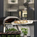 Ooni Koda 12 Outdoor Pizza Oven with a basil topped pizza drawn out on a peel in a cloud of steam on a wooden garden table.