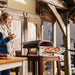 Ooni Koda 12 Outdoor Pizza Oven with a cooked pizza lifted out on a peel while a woman holds a drink beside the table.