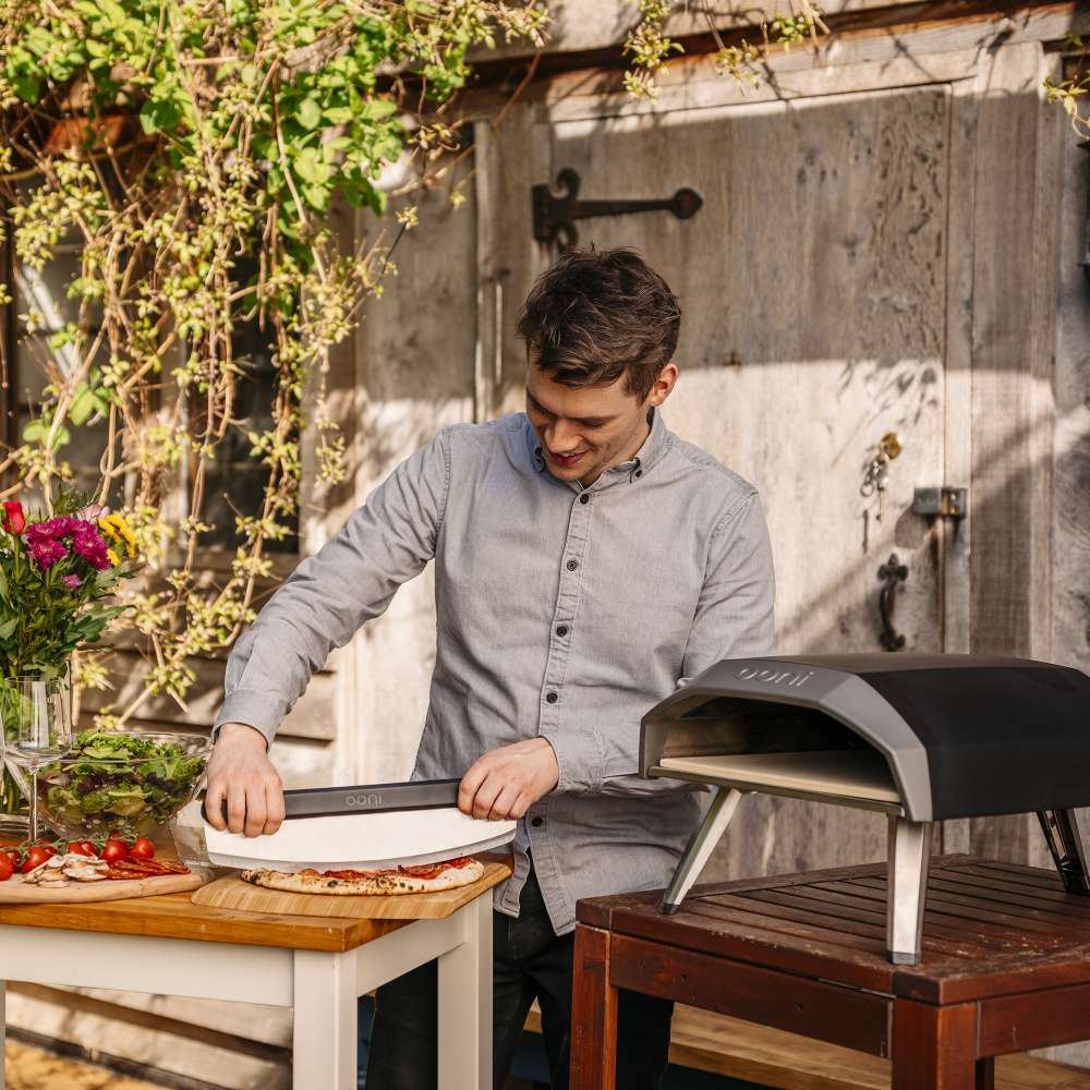 Ooni Koda 12 Outdoor Pizza Oven with a man sliding a pizza on a peel into the lit oven at an outdoor table by a rustic timber wall.