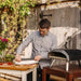 Ooni Koda 12 Outdoor Pizza Oven with a man sliding a pizza on a peel into the lit oven at an outdoor table by a rustic timber wall.