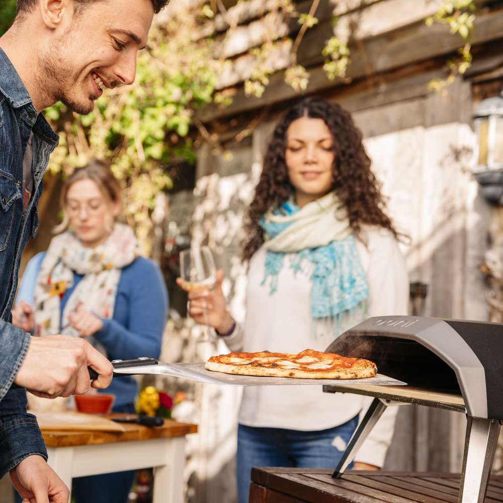 Ooni Koda 12 Outdoor Pizza Oven with friends holding drinks as a man takes a cooked pizza out on a peel.