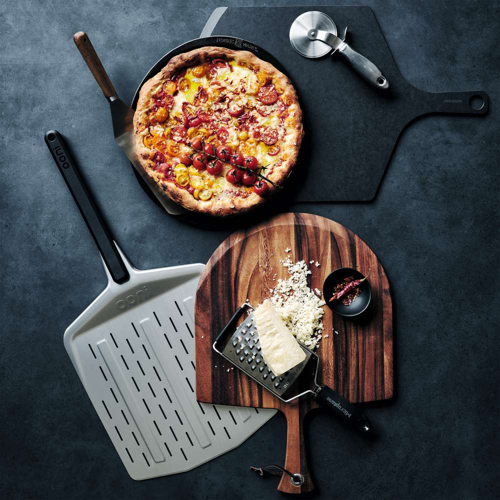 Ooni Perforated Pizza Peel laid on a dark surface beside a cast iron pan with a cooked pizza, a wheel cutter and a board with grated cheese.