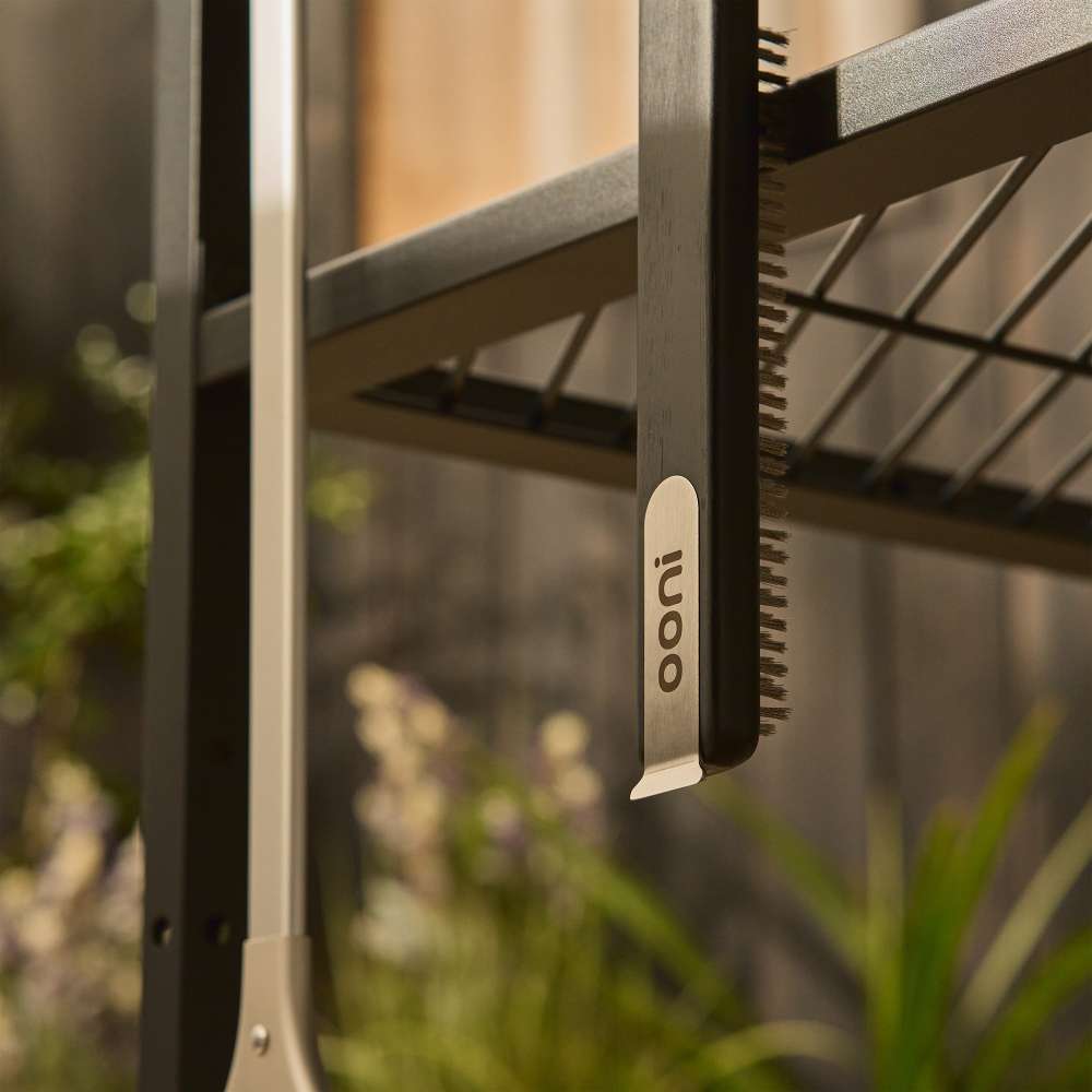 Ooni Pizza Oven Brush hanging by its stainless hook from the rail of an outdoor table with plants blurred behind.