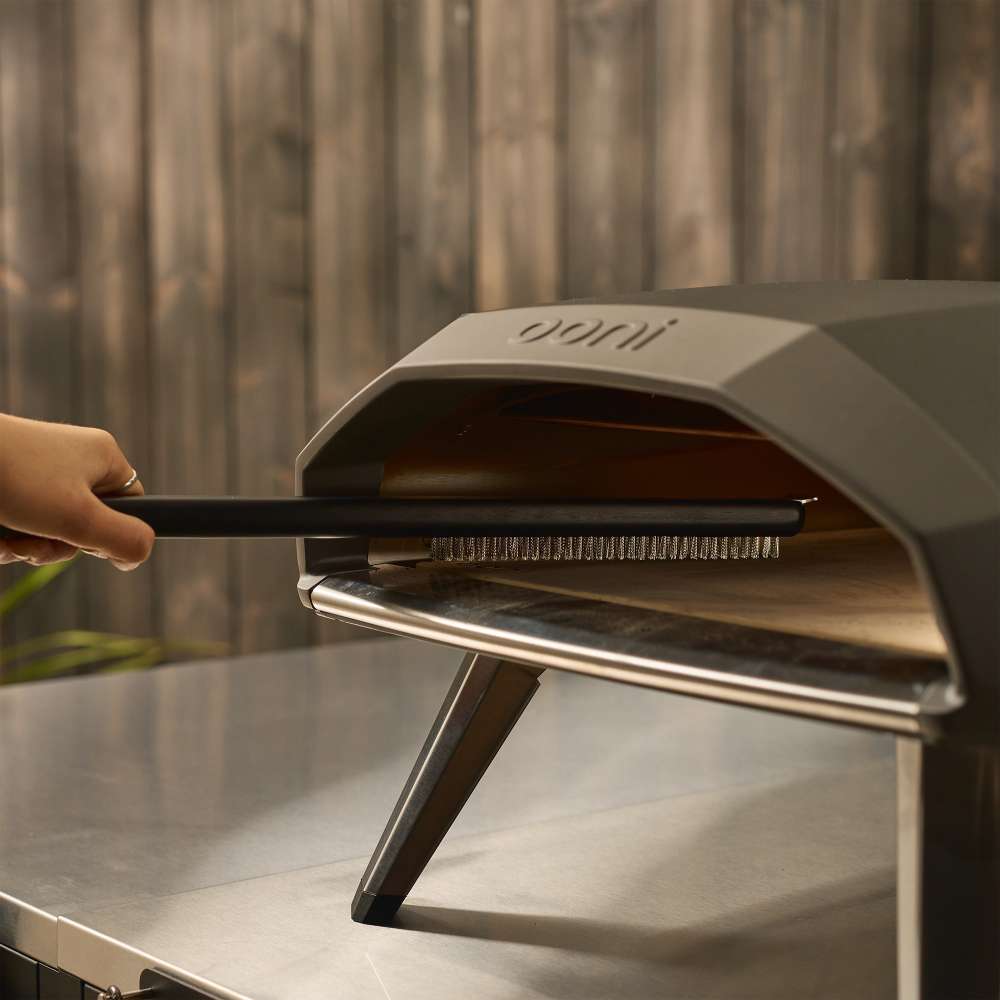 Ooni Pizza Oven Brush held by a hand sweeping inside the mouth of a cream pizza oven on a stainless outdoor table.
