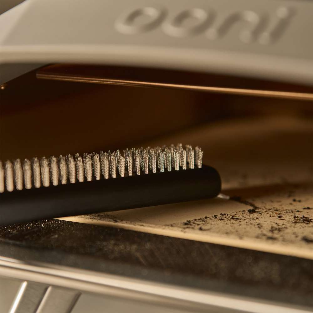 Ooni Pizza Oven Brush resting bristles up on the ash dusted stone at the mouth of a cream pizza oven.