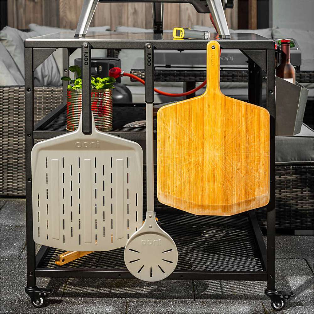 Ooni Pizza Turning Peel hanging on an outdoor table rail beside a perforated pizza peel and a wooden peel with potted basil on the shelf.