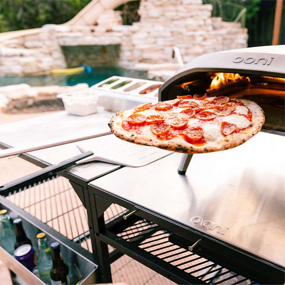 Ooni Pizza Turning Peel holding a cooked tomato and mozzarella pizza at the mouth of a lit oven on an outdoor table beside a pool.