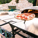 Ooni Pizza Turning Peel holding a cooked tomato and mozzarella pizza at the mouth of a lit oven on an outdoor table beside a pool.