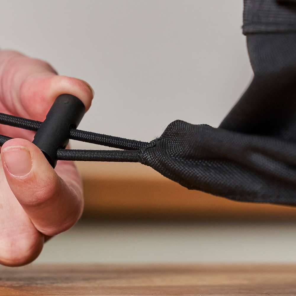 Ooni Volt Pizza Oven Cover with a hand gripping the black cord lock on the elastic hem cord against a pale gray background.