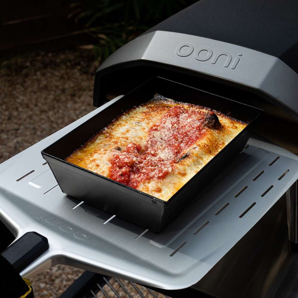 Small Ooni Detroit Pizza Pan holding a baked cheese pizza with stripes of red sauce, sitting on the front ledge of a black Ooni oven outdoors.