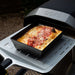 Small Ooni Detroit Pizza Pan holding a baked cheese pizza with stripes of red sauce, sitting on the front ledge of a black Ooni oven outdoors.