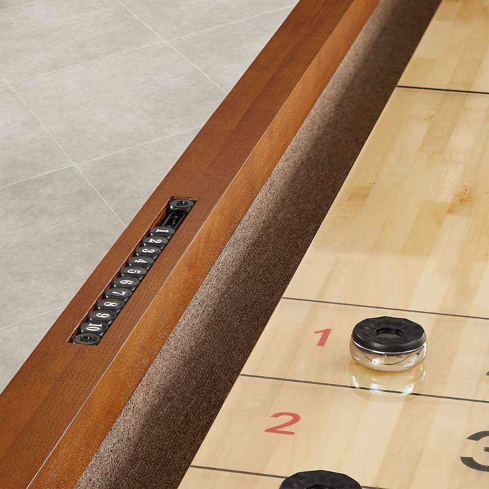 Walnut Crate & Barrel Shuffleboard in close up at the scoring end showing red and black numbers on the pale maple playfield, a black and chrome puck, the brown carpeted alley and a black score counter set into the rail