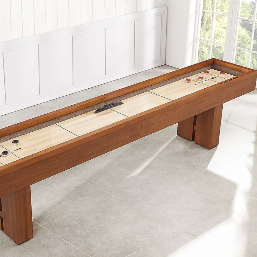 Walnut Crate & Barrel Shuffleboard seen at an angle along its length in a bright room with white panelled walls and a window, with red pucks and the black table brush resting on the pale maple playfield
