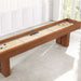 Walnut Crate & Barrel Shuffleboard seen at an angle along its length in a bright room with white panelled walls and a window, with red pucks and the black table brush resting on the pale maple playfield