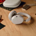 White Sonos Ace Wireless Headphones with Noise Cancellation lying on a wooden desk beside a phone and the closed carrying case