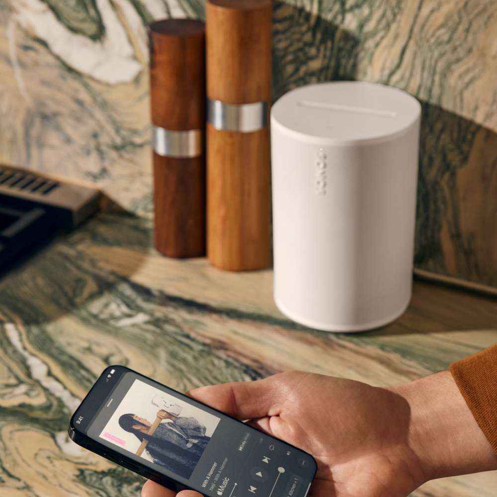 White Sonos Era 100 Next-Gen Smart Speaker on a marble counter beside wooden grinders as a hand holds a phone showing the Sonos app playback screen