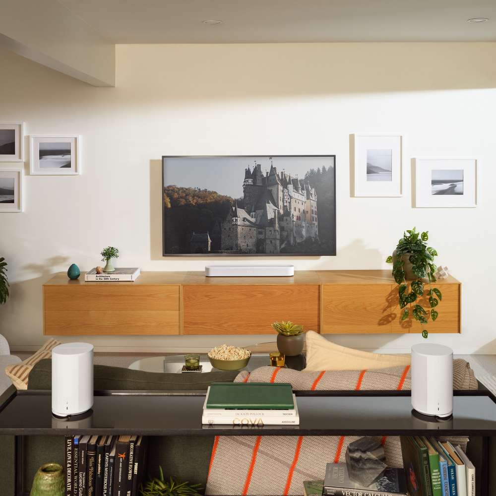 White Sonos Era 100 Next-Gen Smart Speaker two of the speakers on a shelf behind a sofa facing a wall-mounted television above a light wood console in a living room