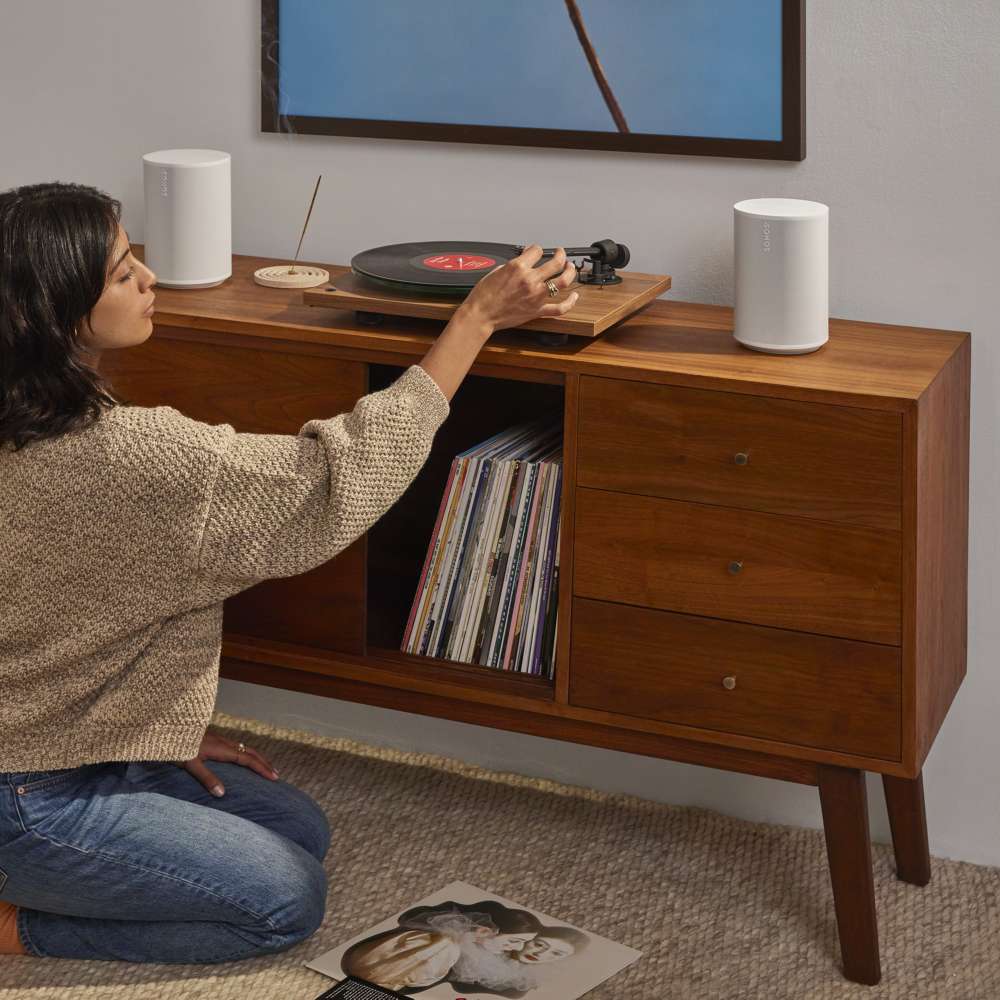 White Sonos Era 100 SL Compact Wireless Speaker a pair of the speakers flanking a record player on a walnut credenza as a woman kneels to pick a vinyl record from the shelf below