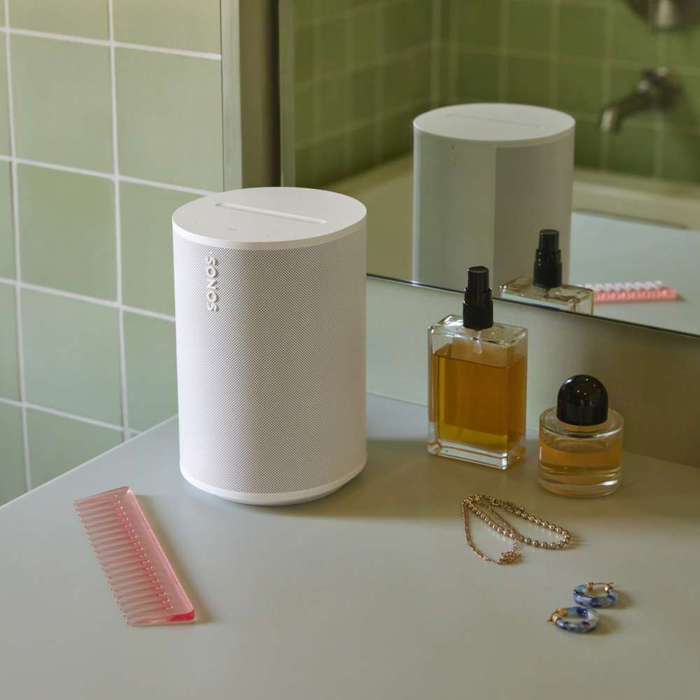 White Sonos Era 100 SL Compact Wireless Speaker standing on a bathroom counter beside perfume bottles, a pink comb and jewellery in front of a green tiled wall