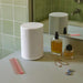 White Sonos Era 100 SL Compact Wireless Speaker standing on a bathroom counter beside perfume bottles, a pink comb and jewellery in front of a green tiled wall