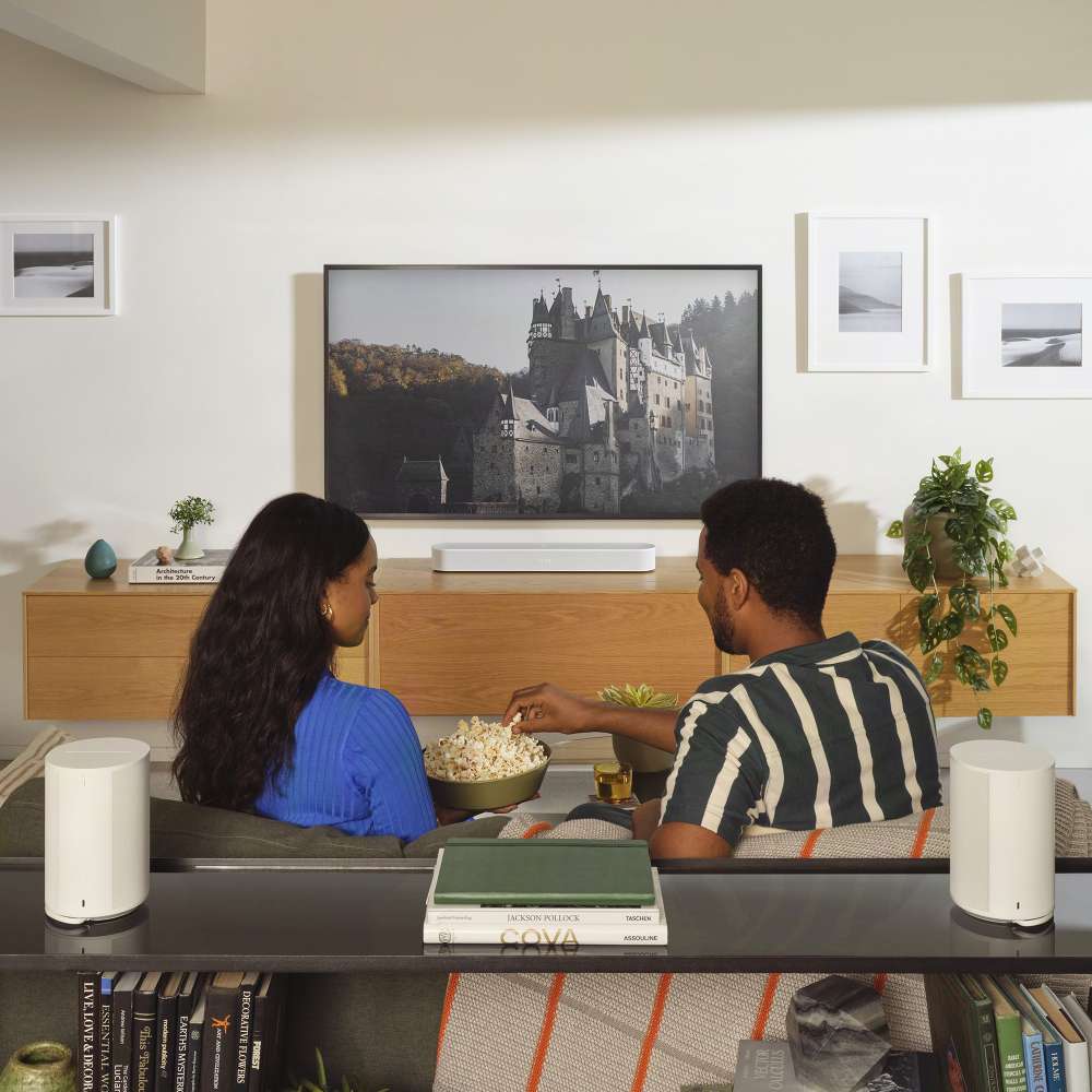 White Sonos Era 100 SL Compact Wireless Speaker two of the speakers on a console behind a couple eating popcorn as they watch a wall-mounted television in a bright living room