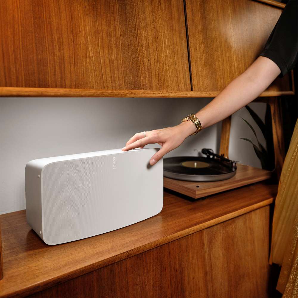 White Sonos Five Speaker a hand placing the speaker horizontally on a wooden shelf next to a record player