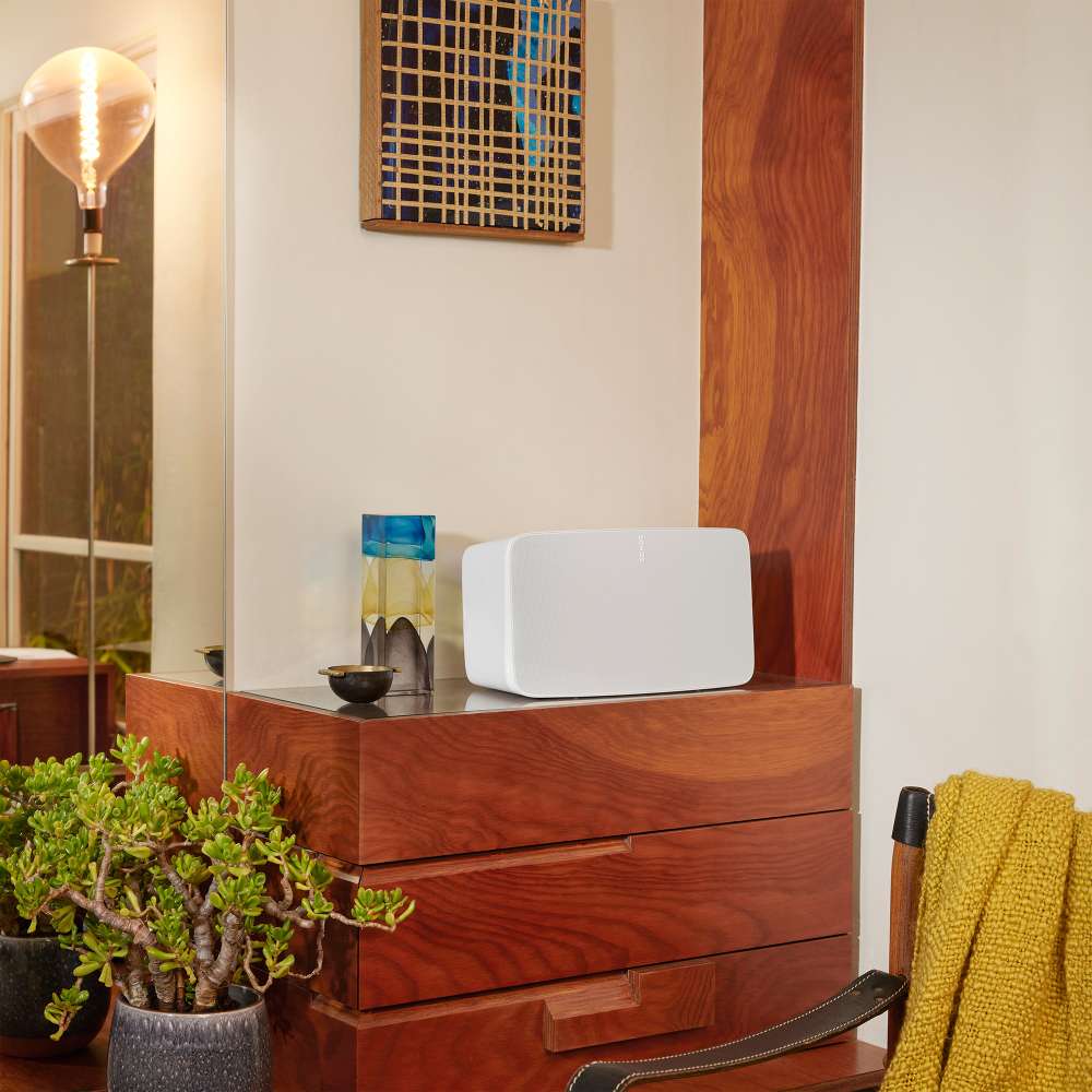 White Sonos Five Speaker sitting horizontally on top of a wooden chest of drawers beside a small vase with a yellow throw and a plant nearby