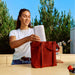 White Sonos Play Portable Bluetooth Speaker being lowered into a red tote bag by a woman standing at an outdoor counter with trees behind her