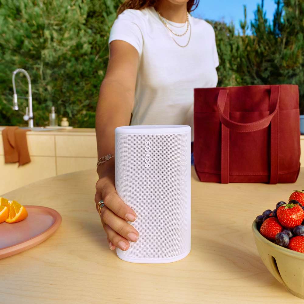 White Sonos Play Portable Bluetooth Speaker held in one hand on an outdoor kitchen counter beside a bowl of strawberries and a red tote bag