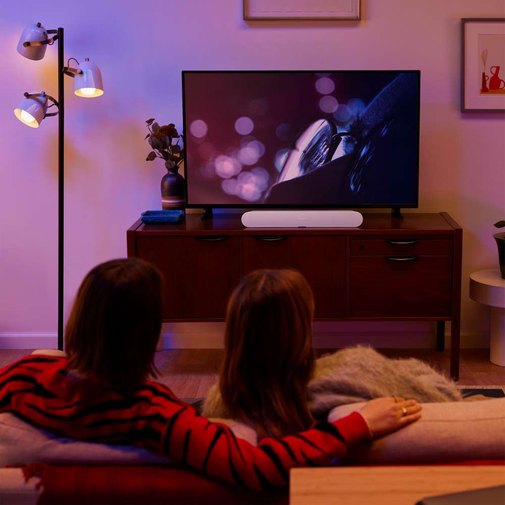White Sonos Ray Sound Bar on a wooden credenza beneath a television as two people watch a film from the sofa in a dimly lit living room