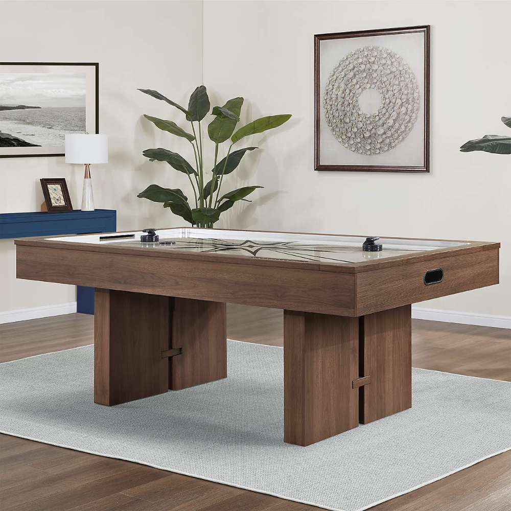Woodgrain Crate & Barrel Air Hockey Table at an angle in a furnished game room on a pale grey rug over a wood floor, with a large plant, a blue sideboard and two framed artworks behind