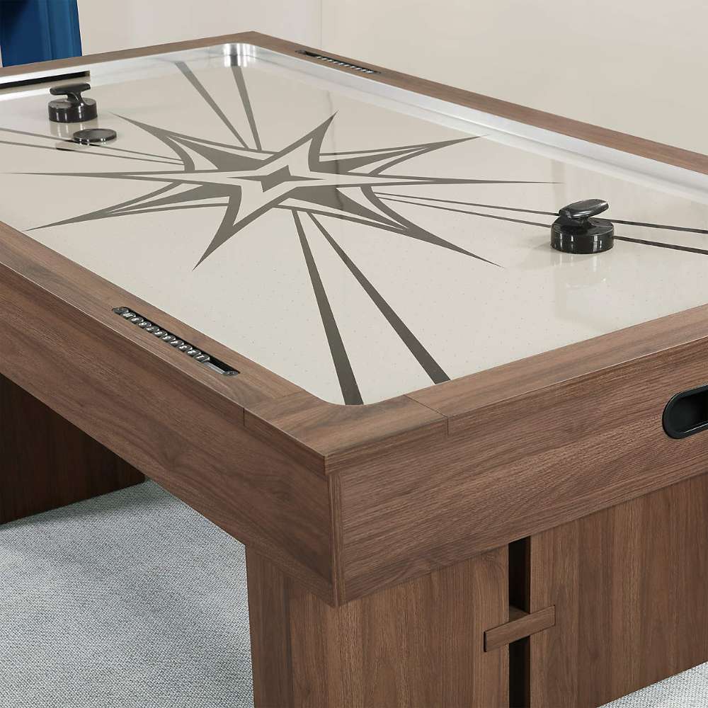 Woodgrain Crate & Barrel Air Hockey Table in close up at one corner showing the cream field with its grey starburst print, two black strikers, a sliding score counter in the rail and the black goal opening