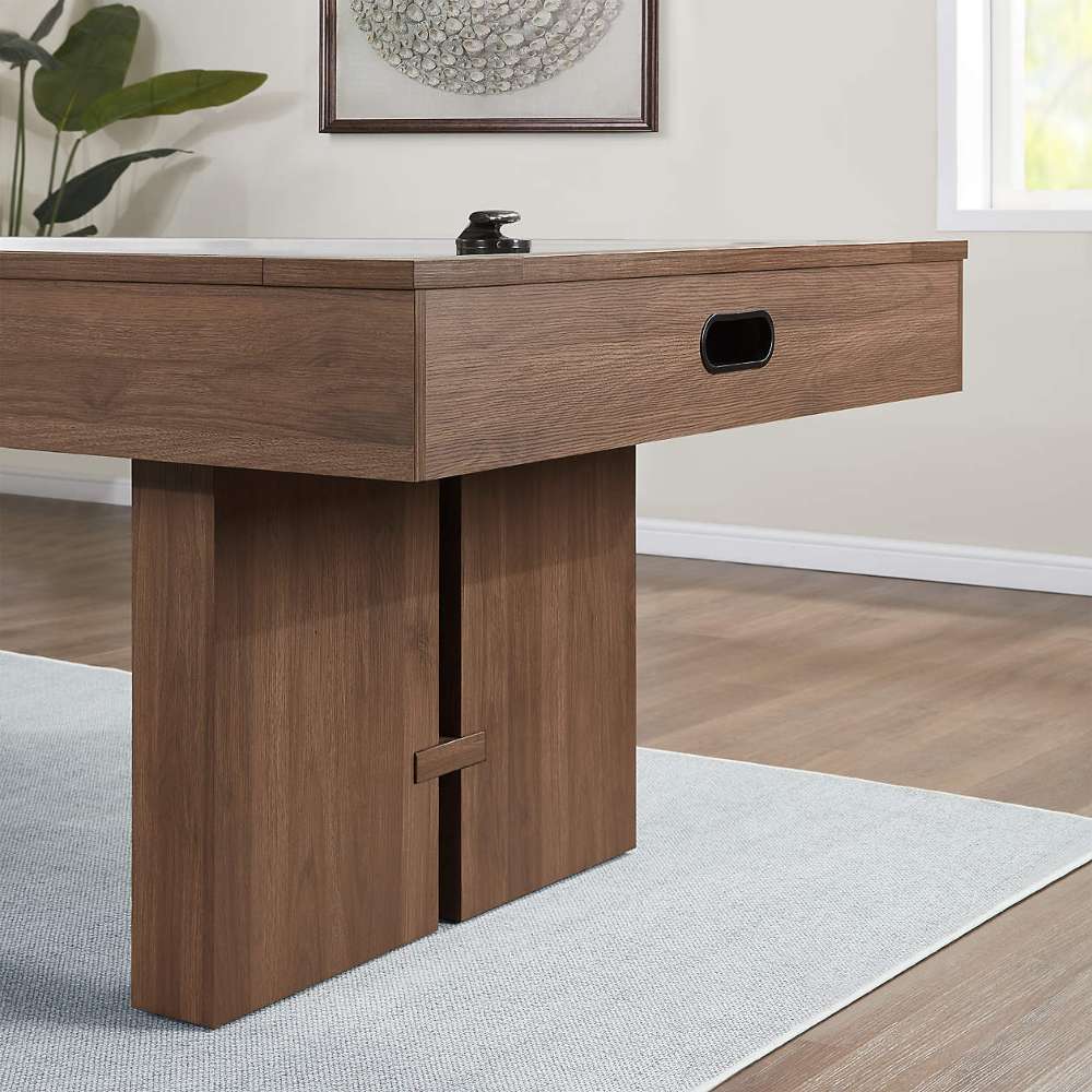 Woodgrain Crate & Barrel Air Hockey Table in close up at one end showing the black oval goal opening in the cabinet and a wide slab leg on a grey rug over a wood floor