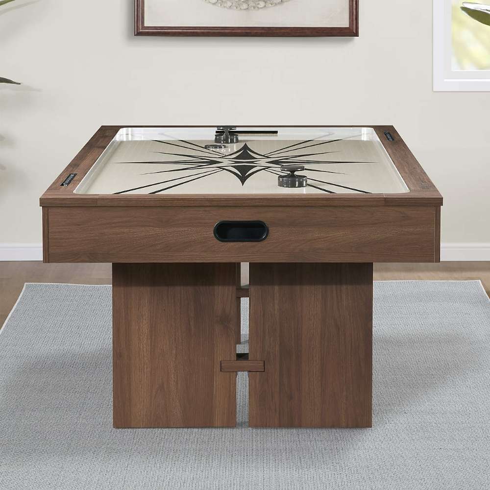 Woodgrain Crate & Barrel Air Hockey Table seen end on in a game room, standing on a pale grey rug against a cream wall with framed art above and showing the starburst field and oval goal opening